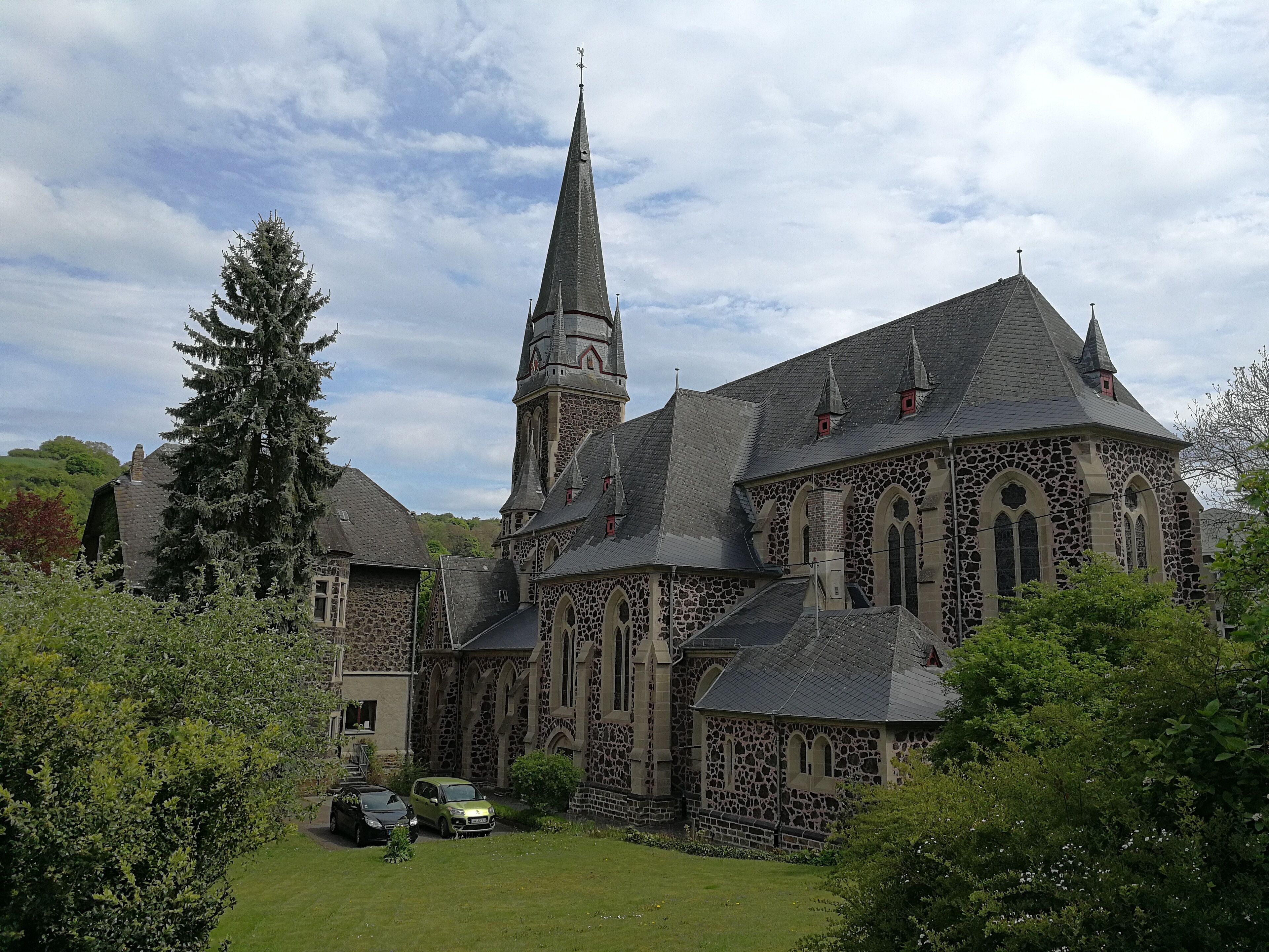 A historic stone church with a tall spire and a smaller tower, surrounded by lush greenery.