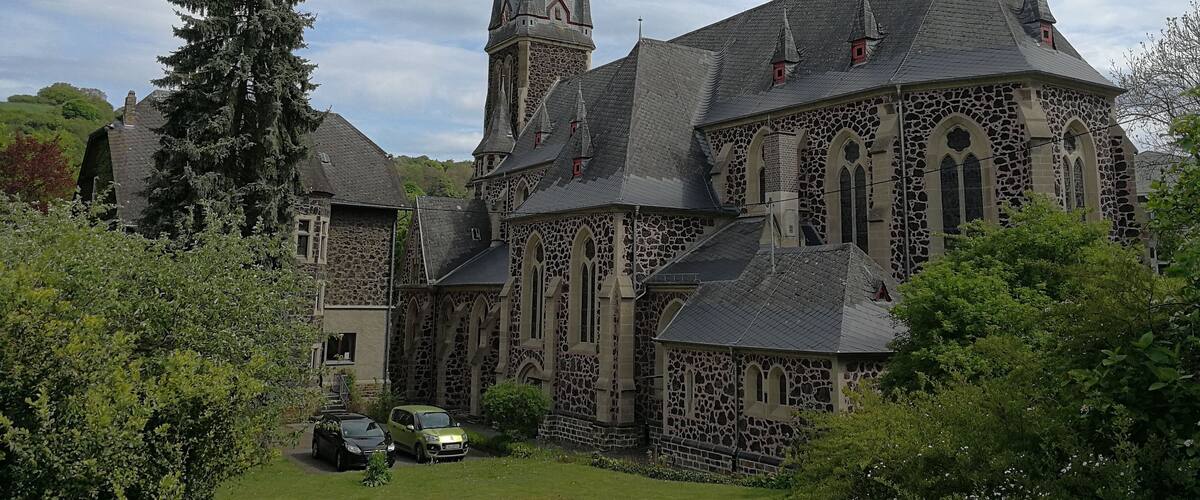 A historic stone church with a tall spire and a smaller tower, surrounded by lush greenery.