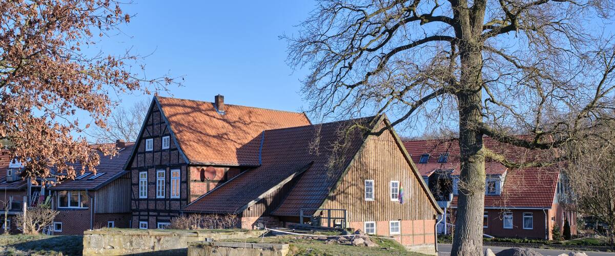 A traditional half-timbered house with a sloped roof, surrounded by a grassy area with rocks and a tree.