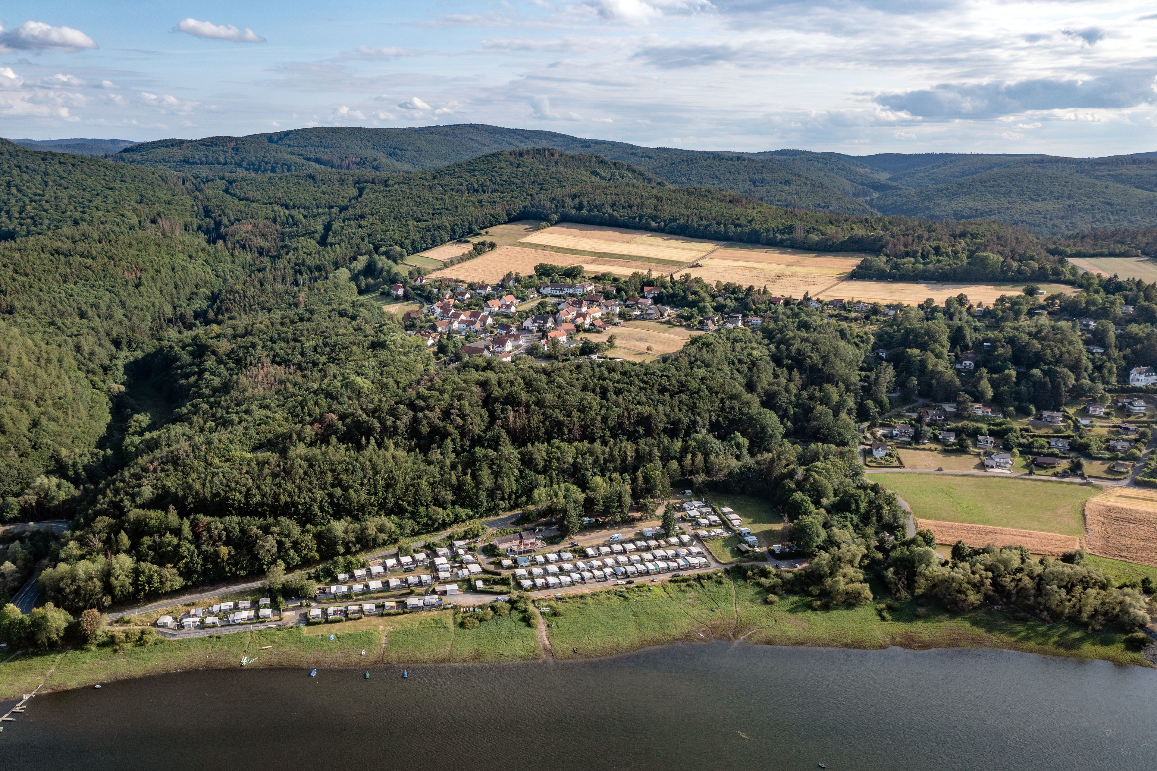 Un terrain de camping au bord d’un lac, avec des emplacements pour camping-cars, entouré de collines boisées et de champs.