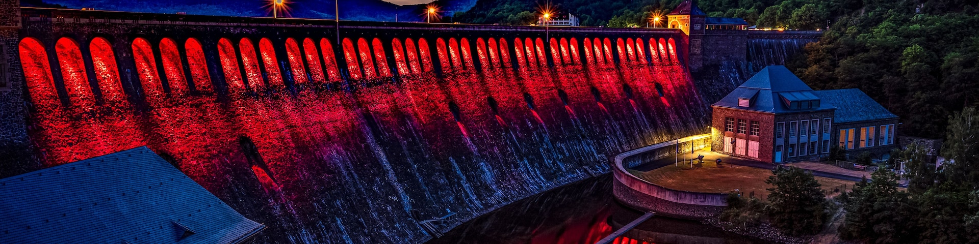 Un barrage illuminé de lumières rouges, une rivière paisible et un paysage montagneux au crépuscule.