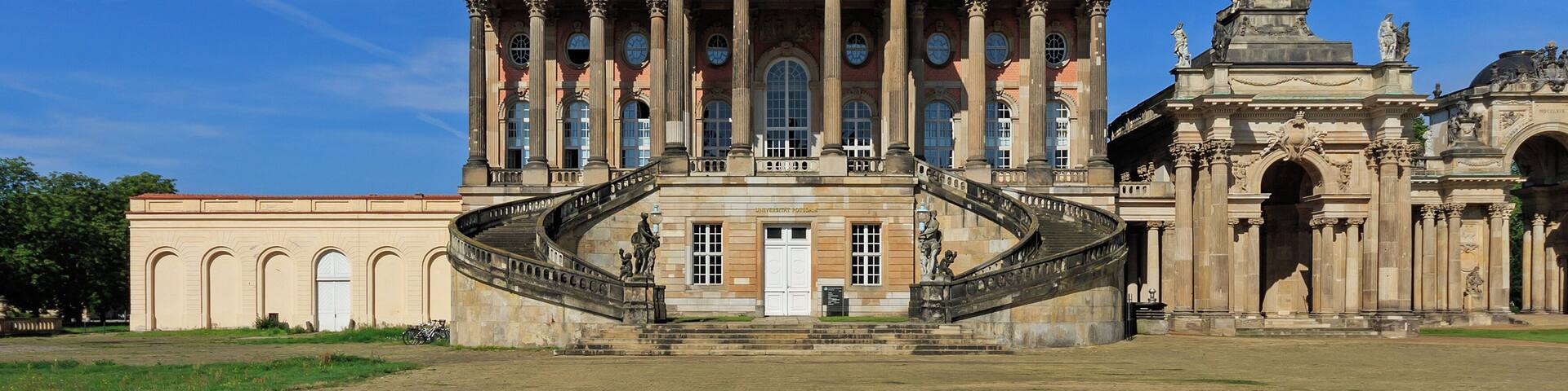 A grand neoclassical building with a central dome, a green spire, and a staircase leading to the entrance.