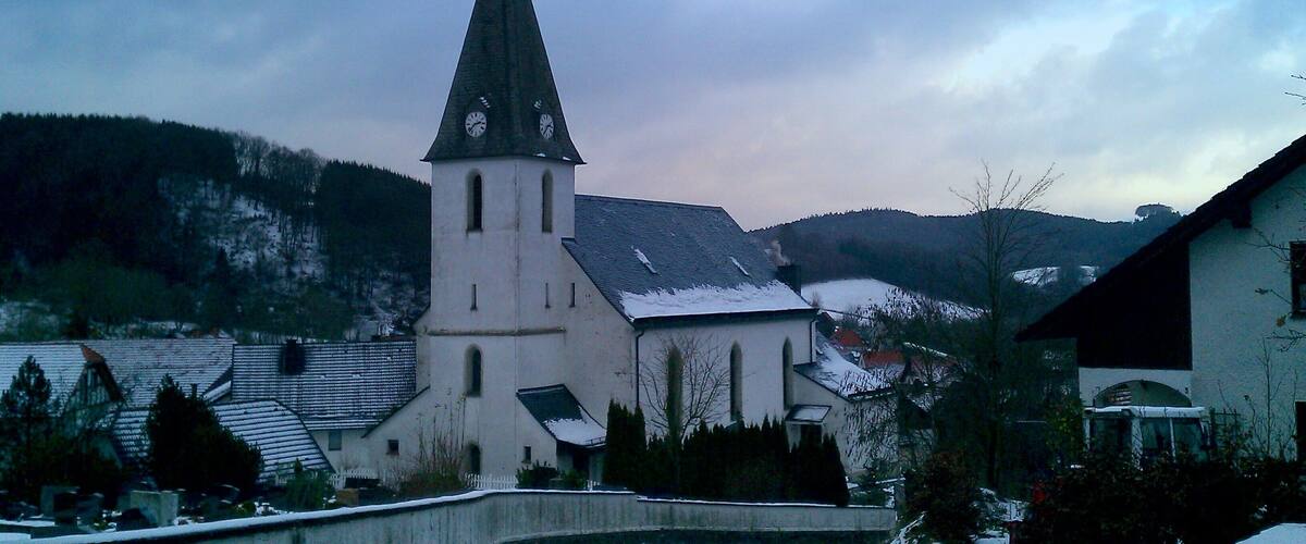 A church with a tall steeple and a clock, surrounded by snow-covered buildings and hills.