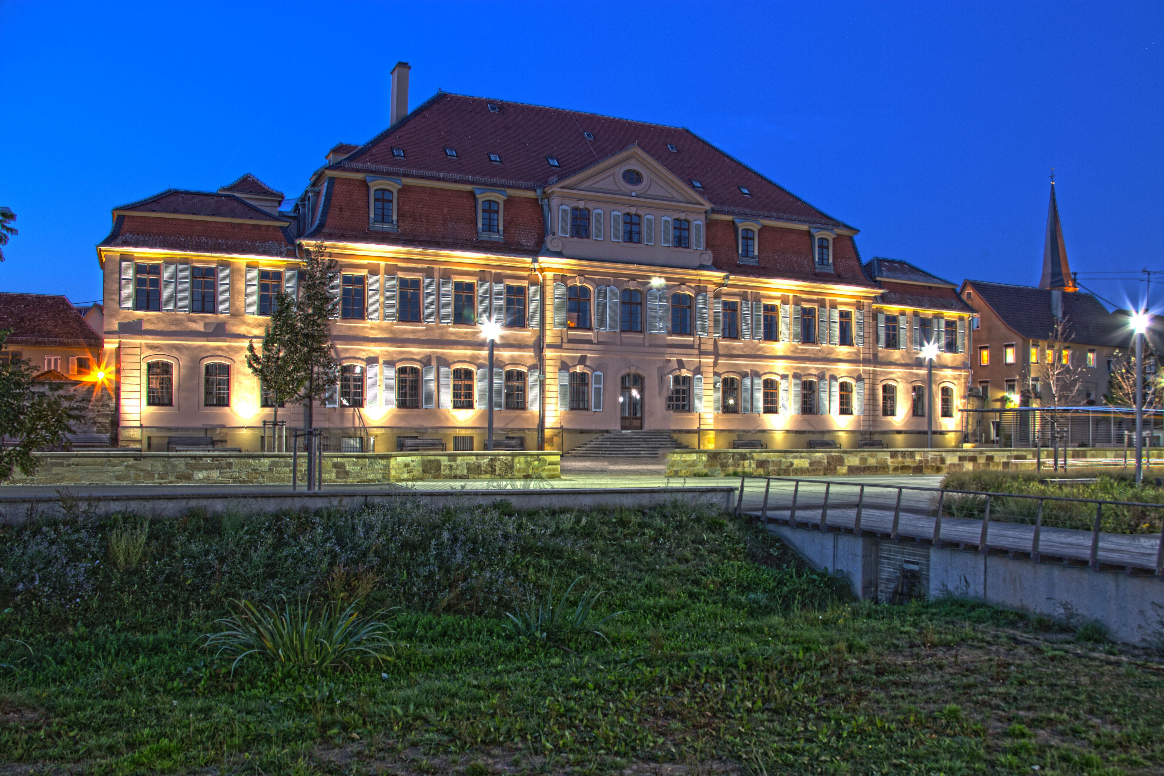 A large, illuminated historic building with a red roof and multiple windows, situated by a canal.