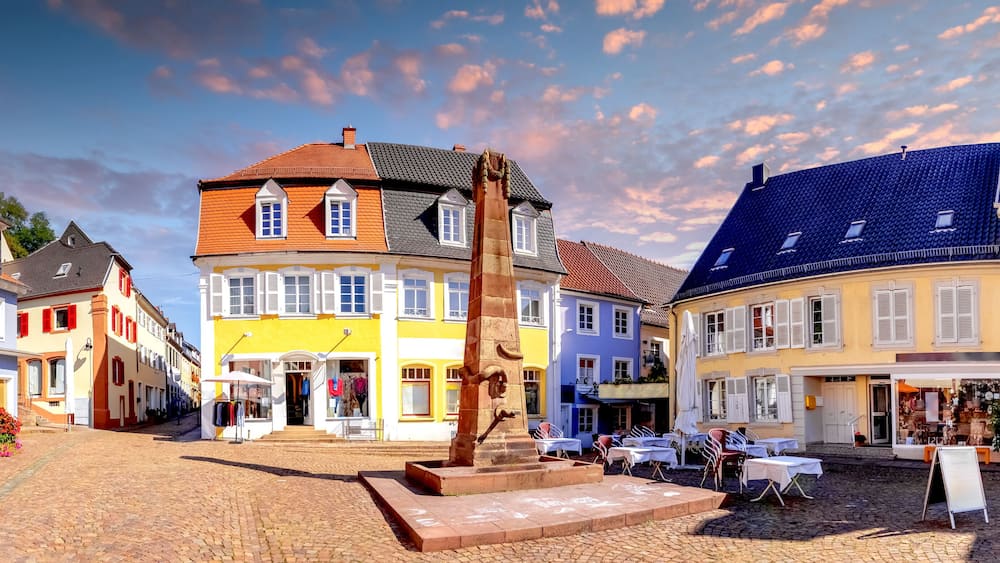 A cobblestone street lined with colorful buildings, a central stone monument, and outdoor seating areas.