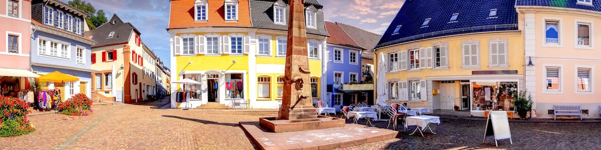 A cobblestone street lined with colorful buildings, a central stone monument, and outdoor seating areas.