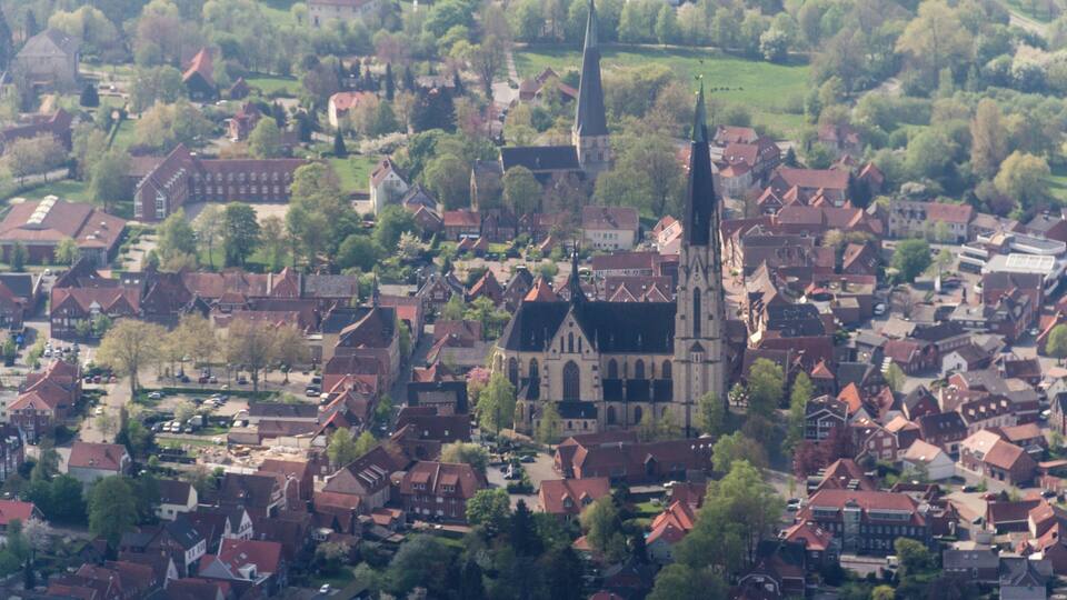 An aerial view of a town with a prominent church with a tall spire, surrounded by red-roofed buildings and green spaces.