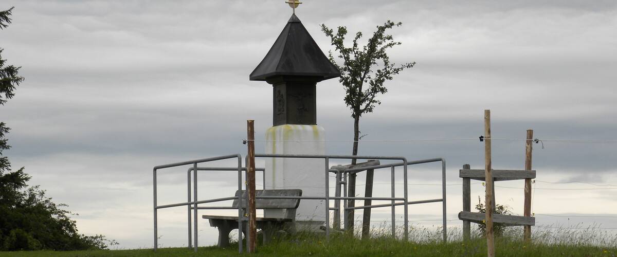 A small tower with a pointed roof and a cross on top, surrounded by a metal fence and a bench.