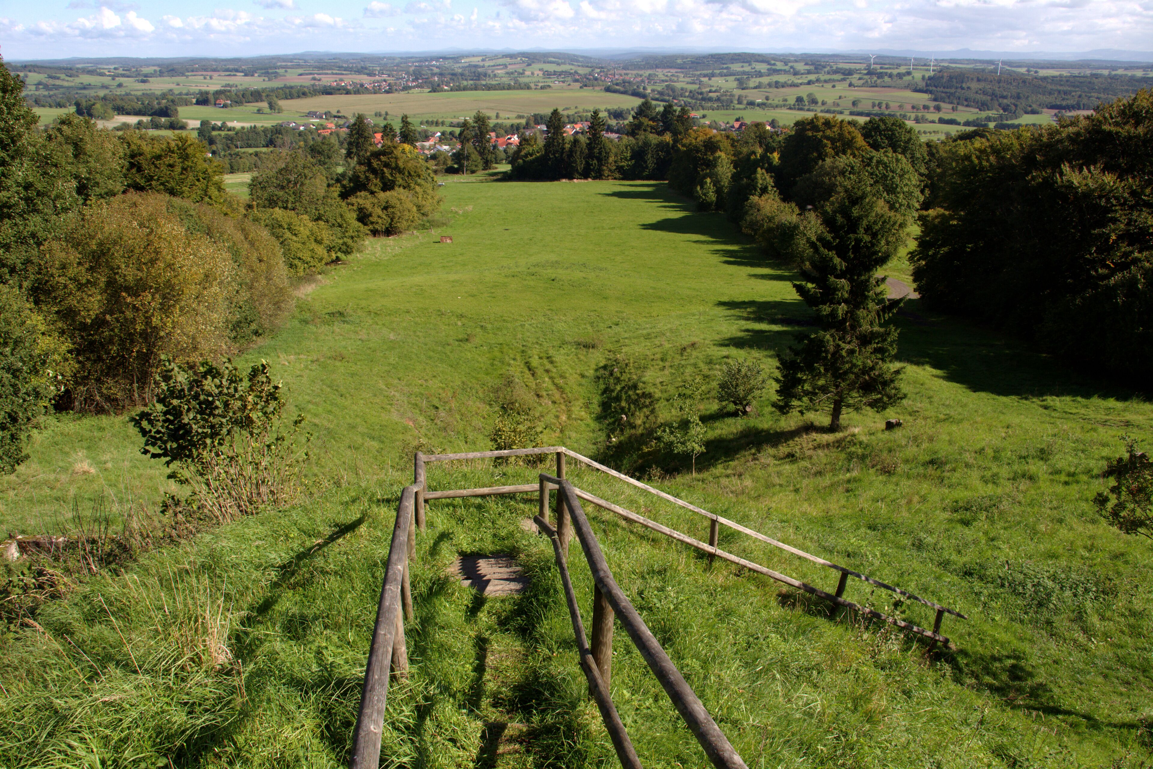 A grassy field with a wooden structure in the foreground and a distant village.