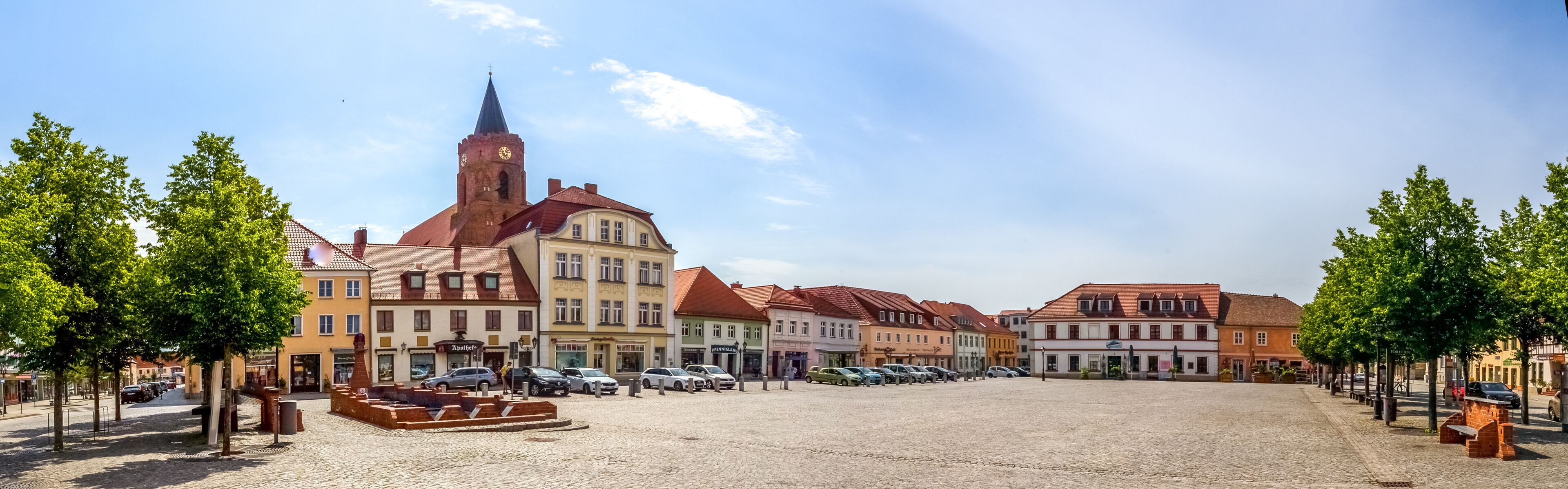 A cobblestone square with historic buildings, a church tower, and parked cars.