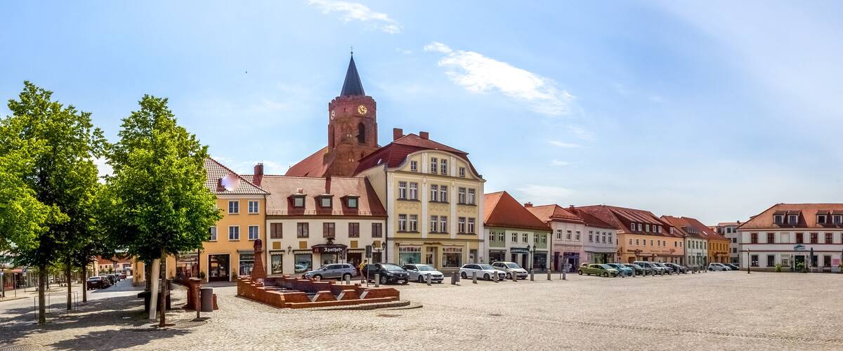 A cobblestone square with historic buildings, a church tower, and parked cars.