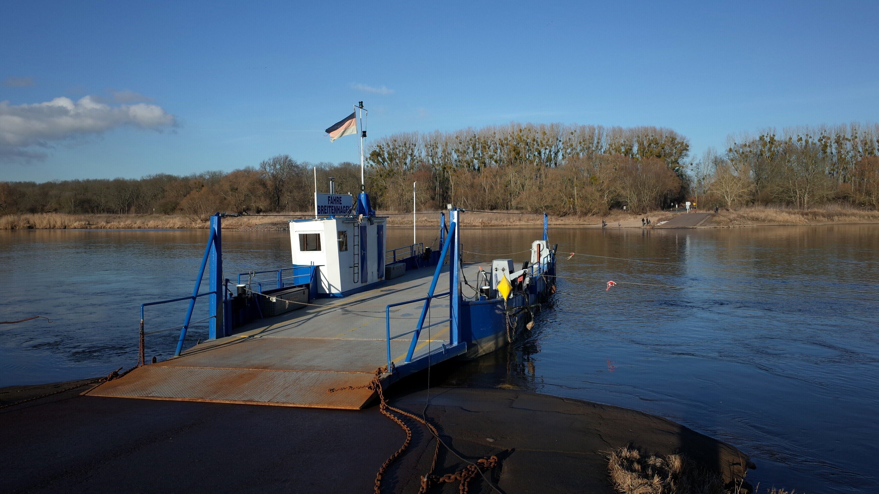 A ferry dock with a blue and white structure, a flag, and a calm river with trees in the background.