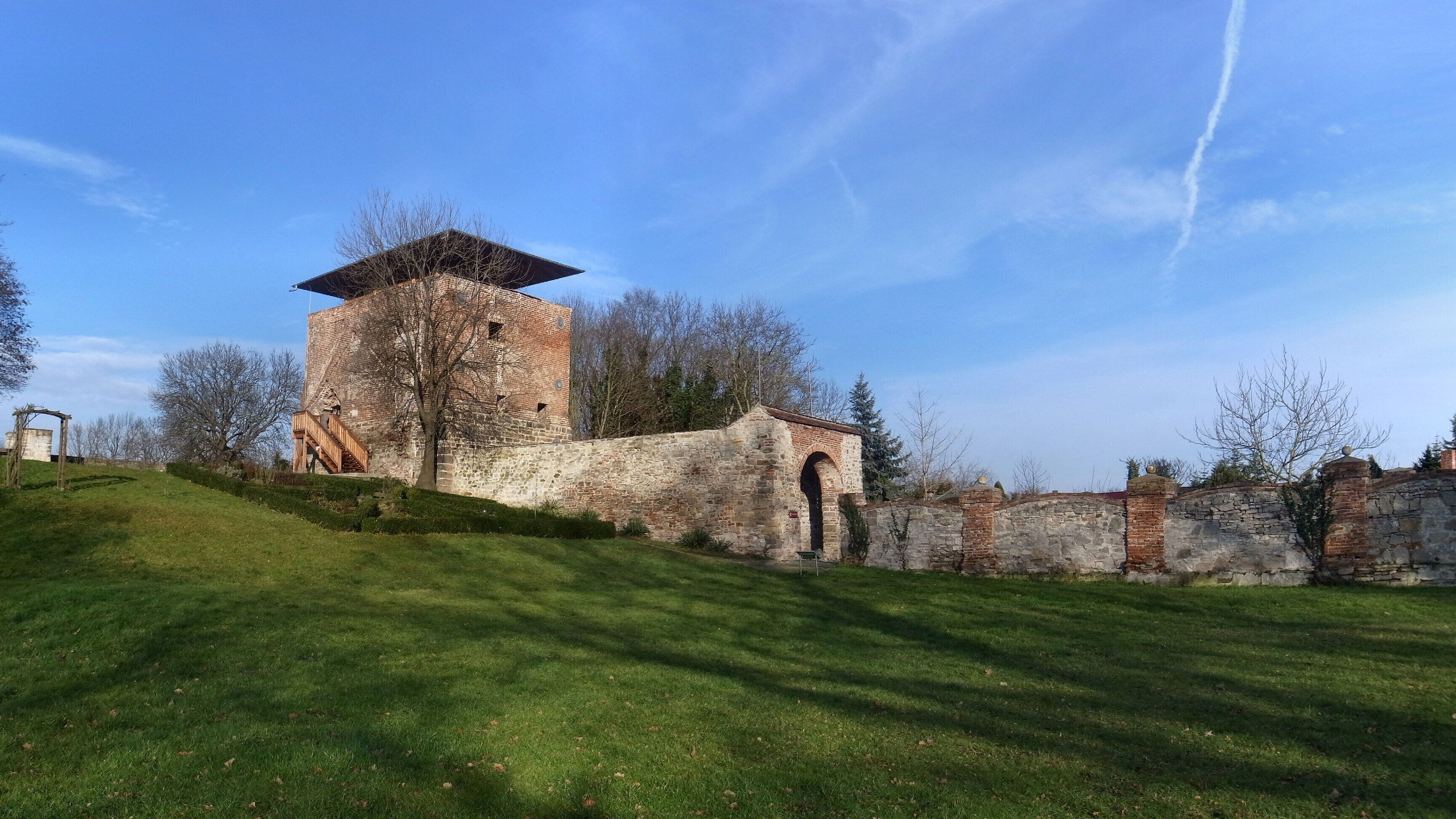 A historic stone tower with a wooden staircase, surrounded by a grassy hill and trees.