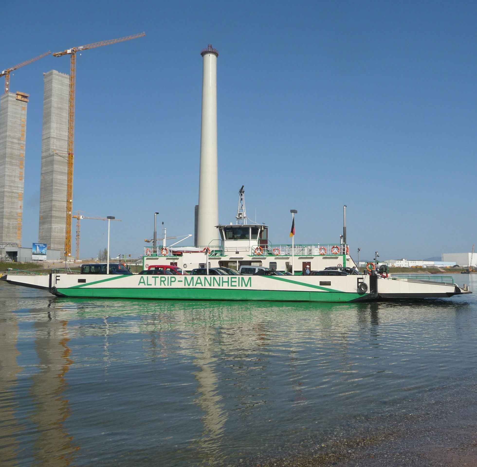 A ferry named ALTRIP-MANNHEIM docked at a port with industrial buildings and cranes in the background.