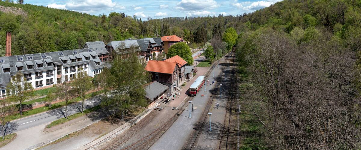 A railway station with a red train, surrounded by buildings and a forested hillside.