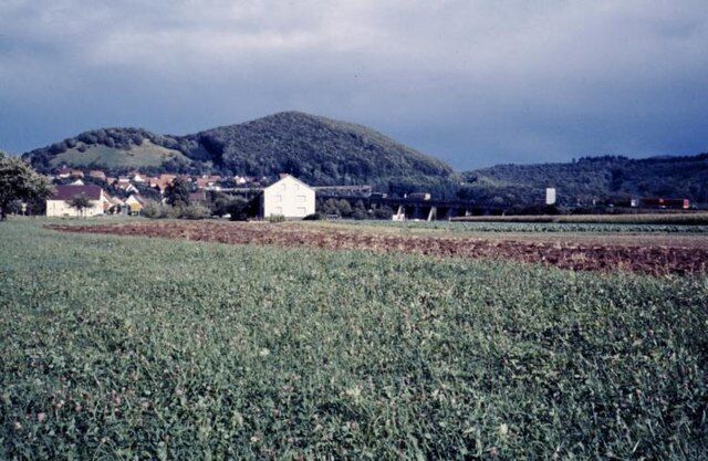 A rural landscape with a field, a white building, and a hill in the background.