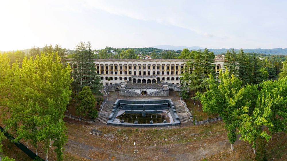 A large, historic building with a central courtyard, surrounded by lush green trees and hills in the background.