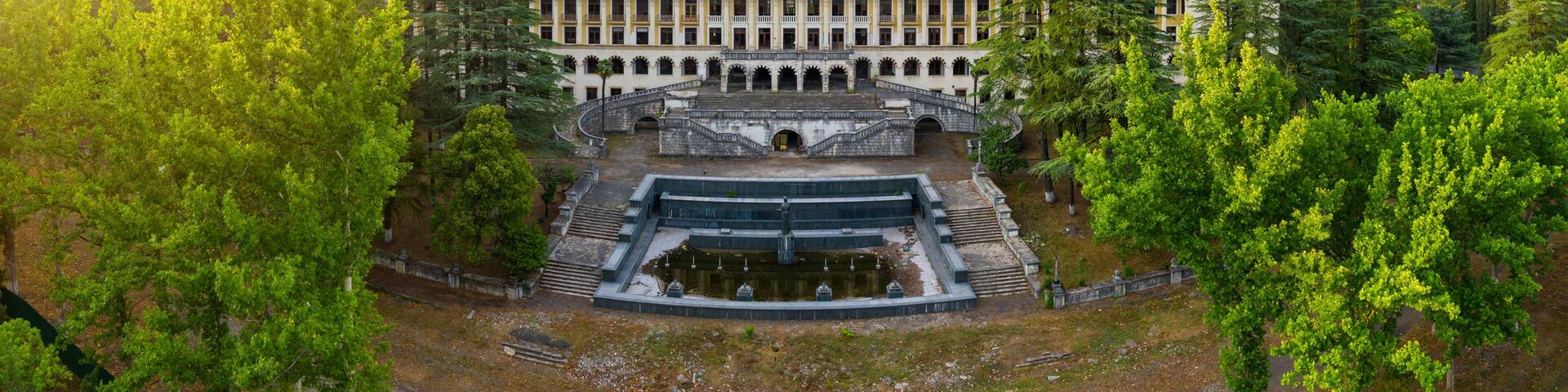 A large, historic building with a central courtyard, surrounded by lush green trees and hills in the background.
