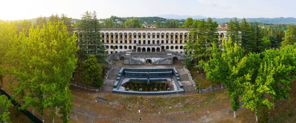 A large, historic building with a central courtyard, surrounded by lush green trees and hills in the background.