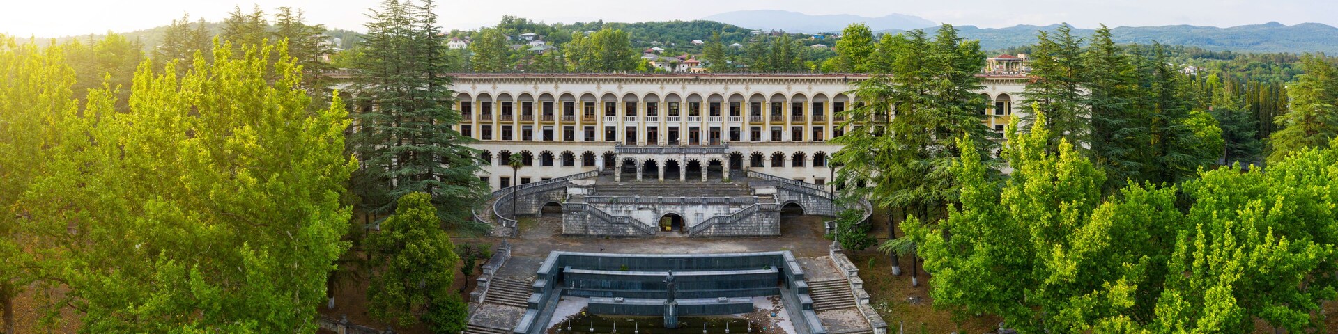A large, historic building with a central courtyard, surrounded by lush green trees and hills in the background.