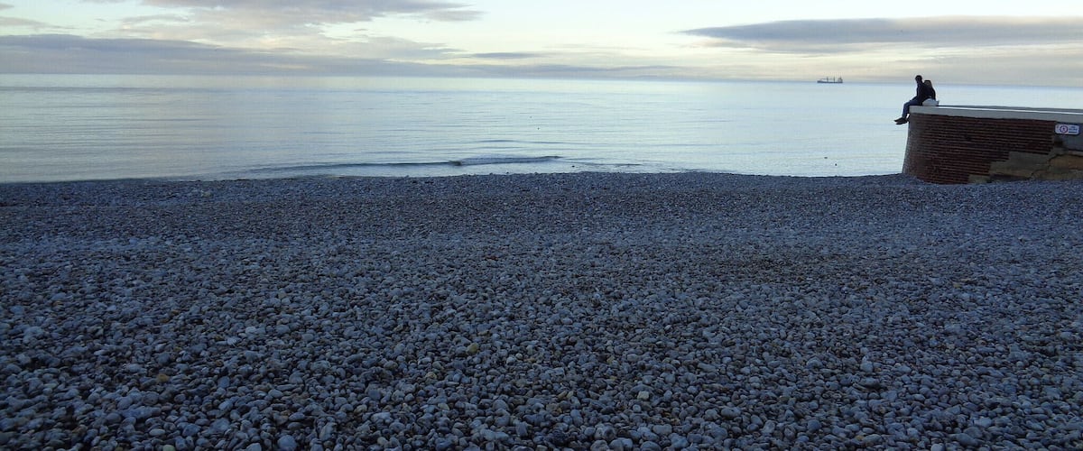 A pebbled beach with a person sitting on a low wall, overlooking calm waters and a distant ship.