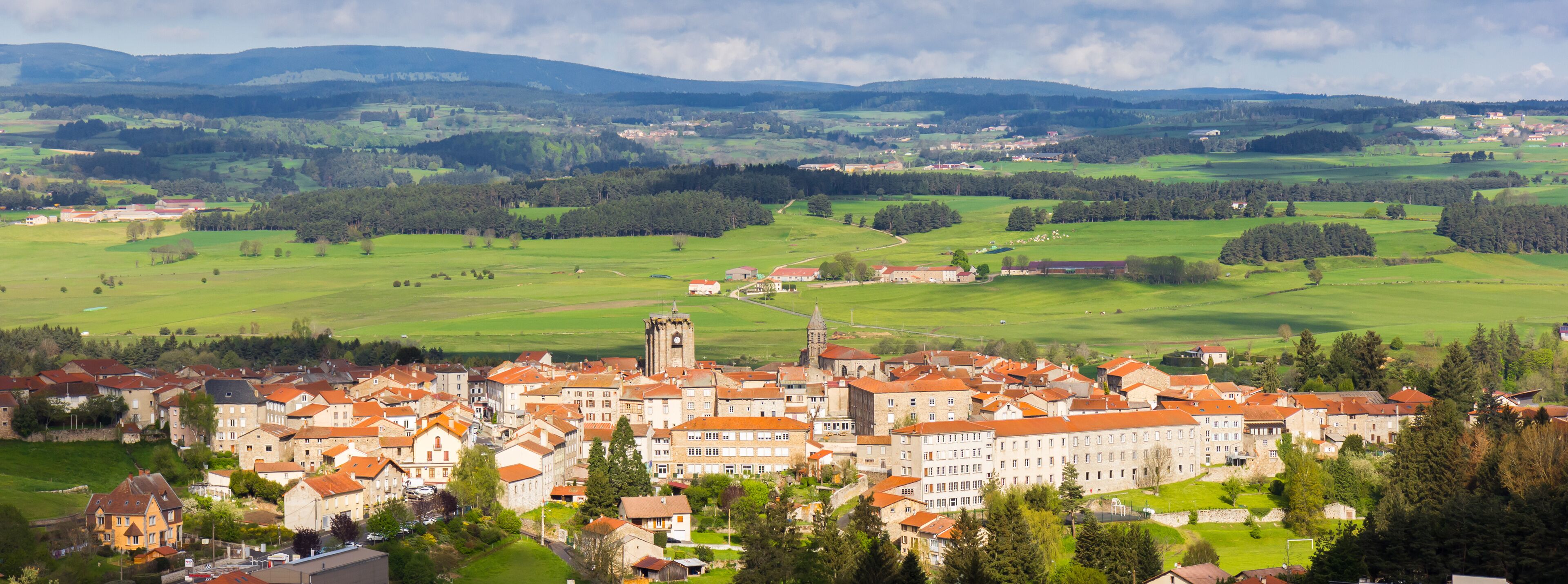 A town with red-roofed buildings, a prominent church tower, and rolling green hills in the background.
