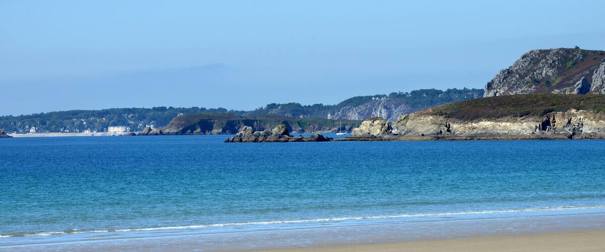 A coastal landscape with a sandy beach, calm sea, rocky cliffs, and a distant hill with sparse vegetation.