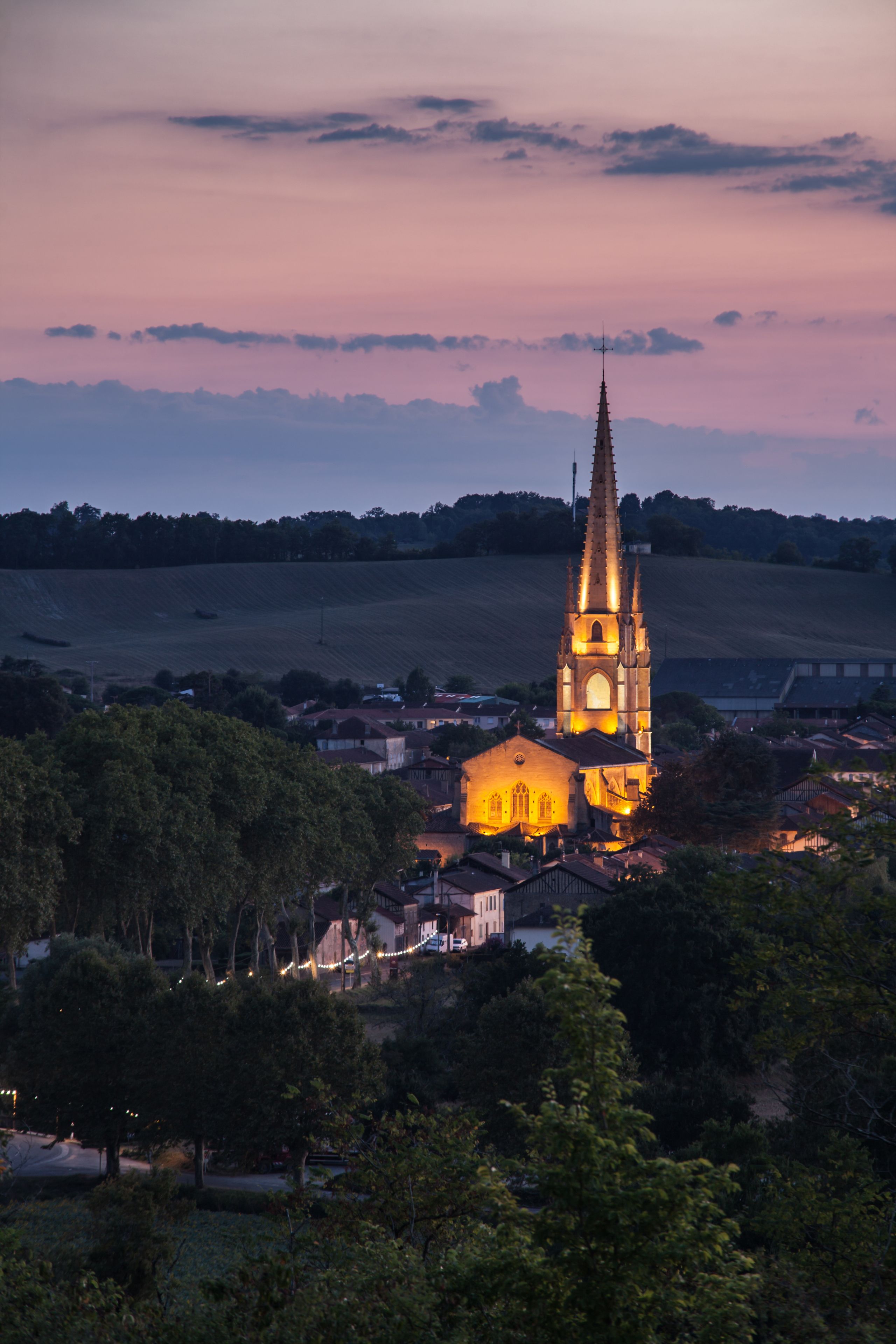 A church with a tall spire, surrounded by trees and buildings, during twilight.