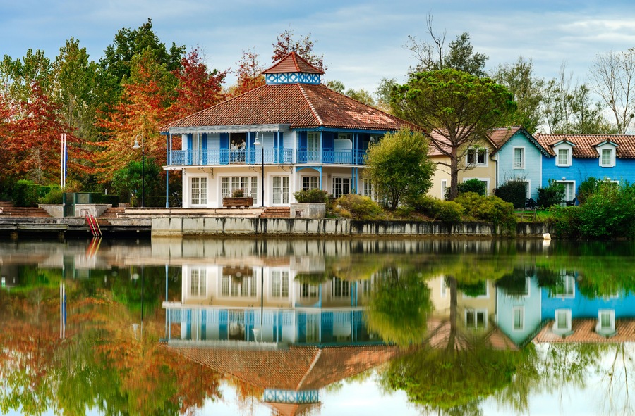 Ein blaues Haus mit einem Ziegeldach, ein Teich, der das Gebäude spiegelt, und im Hintergrund herbstlich gefärbte Bäume.