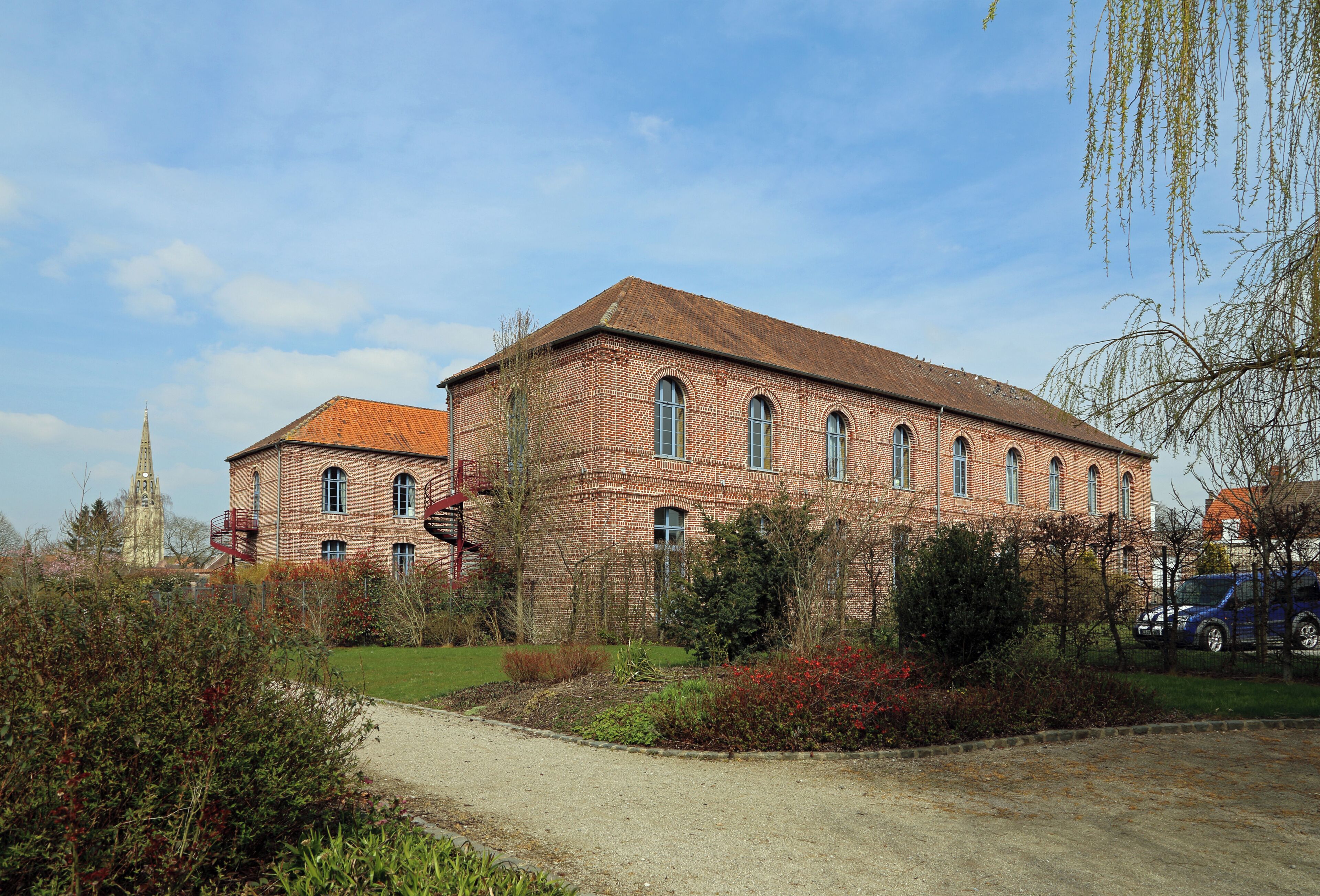 A large brick building with arched windows and a red-tiled roof, surrounded by a landscaped garden with various shrubs and a gravel path.