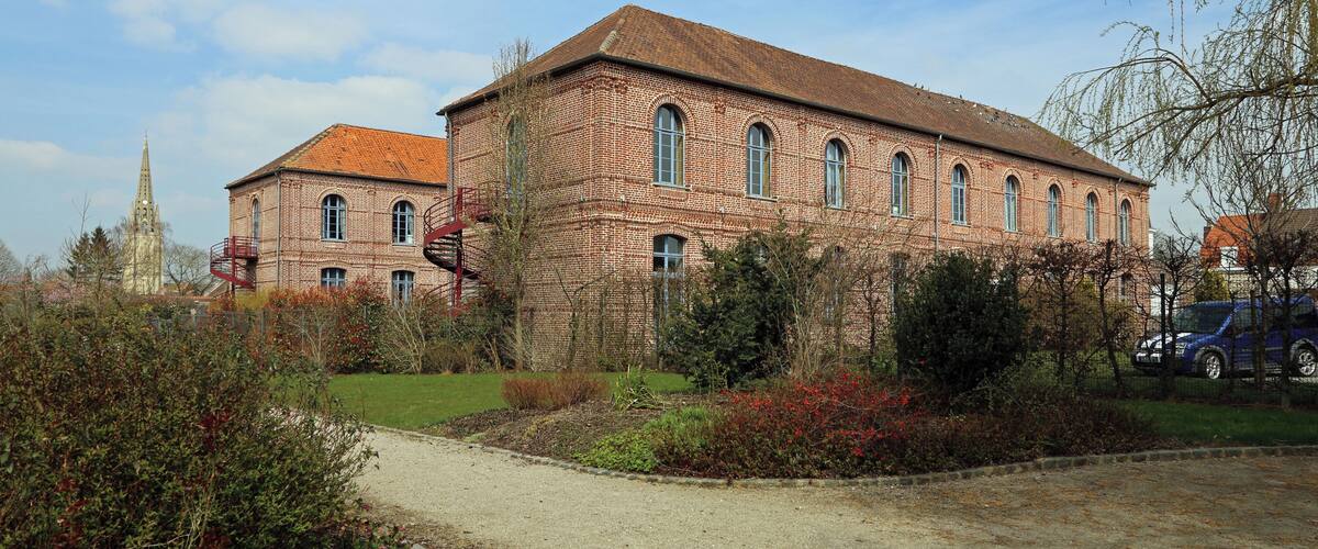A large brick building with arched windows and a red-tiled roof, surrounded by a landscaped garden with various shrubs and a gravel path.