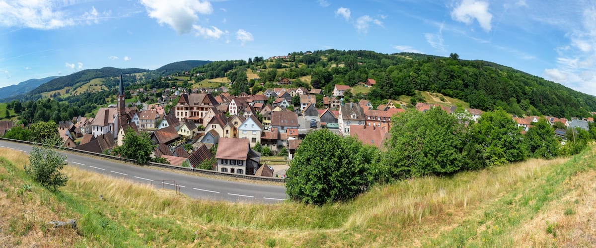 A picturesque village nestled in a valley with a prominent church tower, surrounded by lush green hills and a clear blue sky.