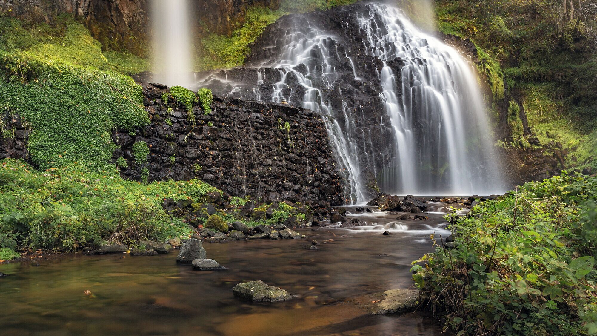 A waterfall cascading over a rocky cliff into a stream surrounded by lush greenery.
