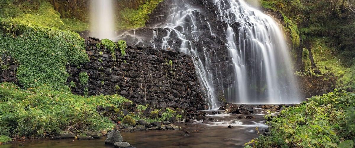 A waterfall cascading over a rocky cliff into a stream surrounded by lush greenery.