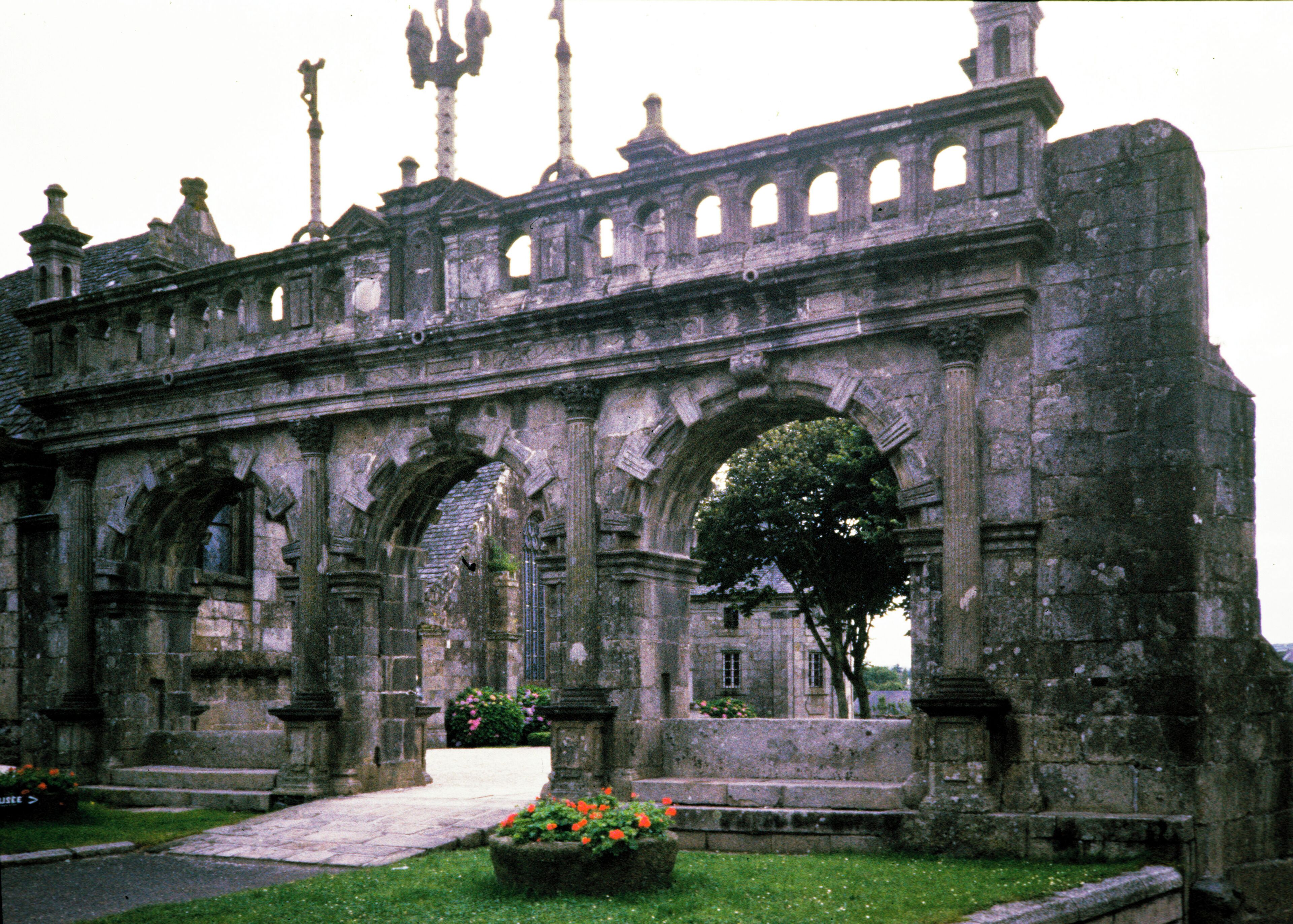 An ancient stone archway with intricate carvings and a garden in the foreground.