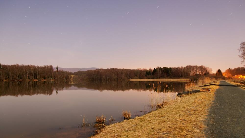 Ein ruhiger See im Abendlicht mit klarem Himmel, einem Weg am Ufer und weiter Ferne Bäume.