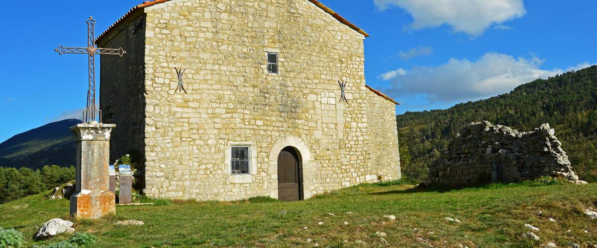 A stone church with a bell tower and a cross, surrounded by a grassy field and mountains.