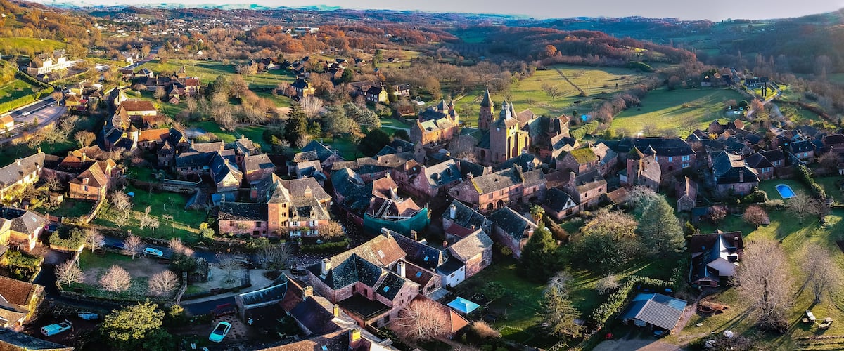 A historic village with medieval architecture, including a church with a tall spire, surrounded by green fields and residential houses.
