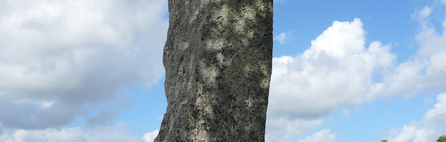 A tall standing stone in a grassy field with a blue sky and clouds.