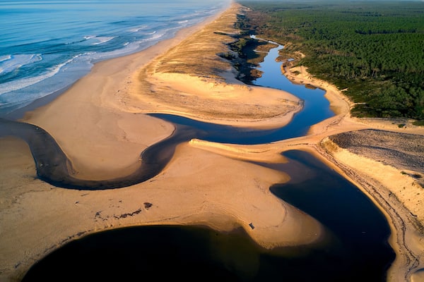 Vue aérienne d’un paysage côtier comprenant une plage de sable, une rivière et une zone boisée.