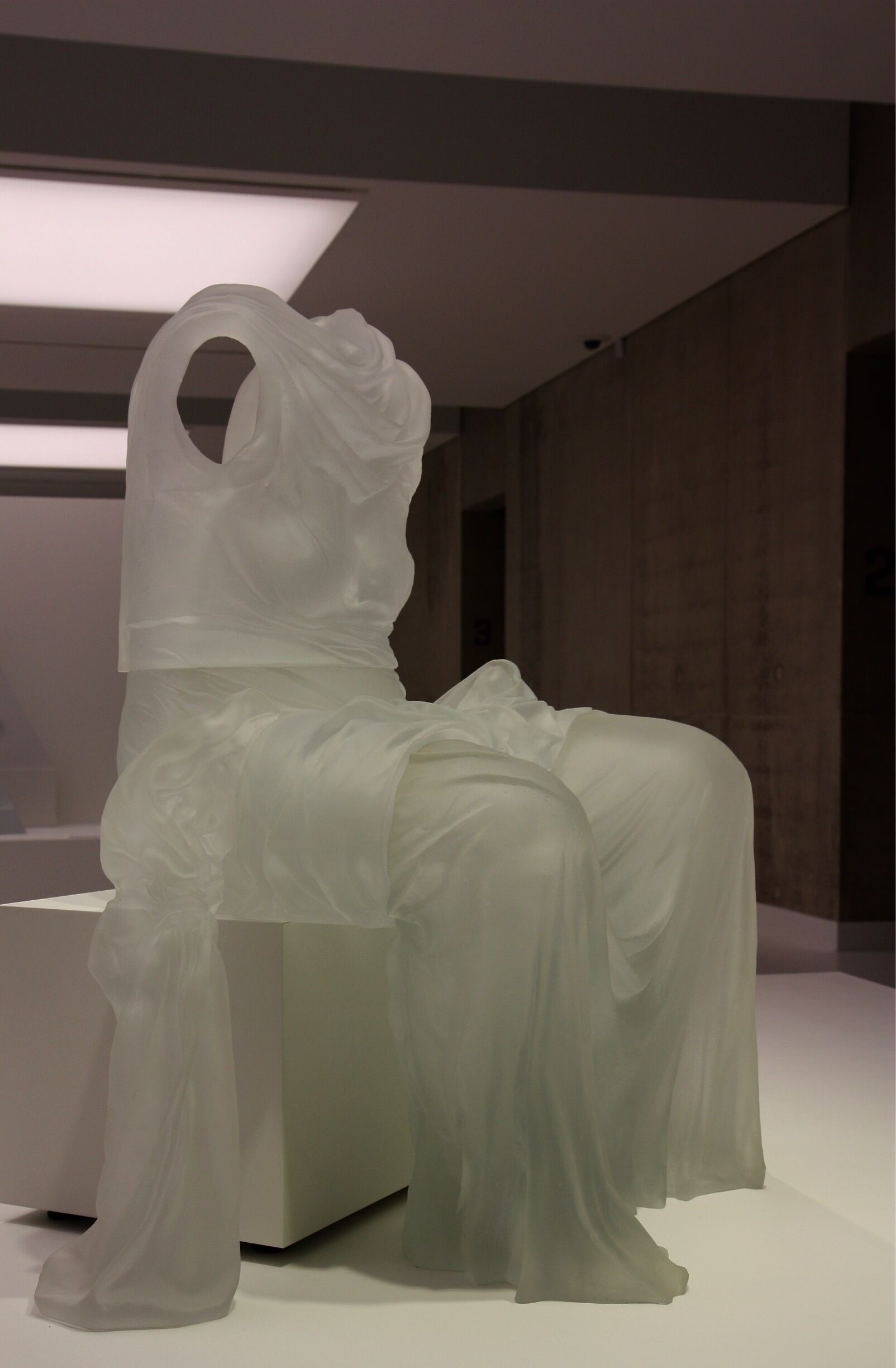 A modern sculpture of a figure in white, seated on a white pedestal.