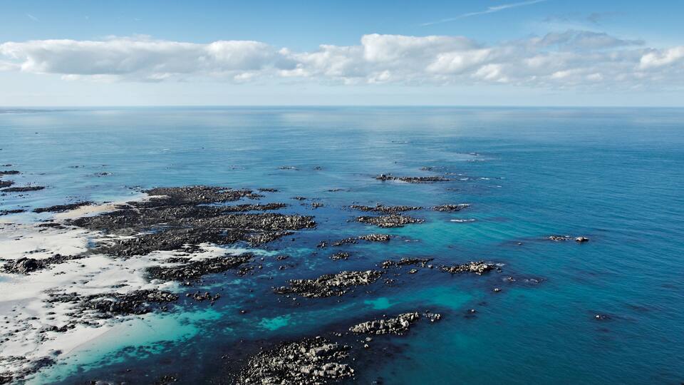 A coastal landscape with clear blue waters, rocky formations, and a sandy beach.