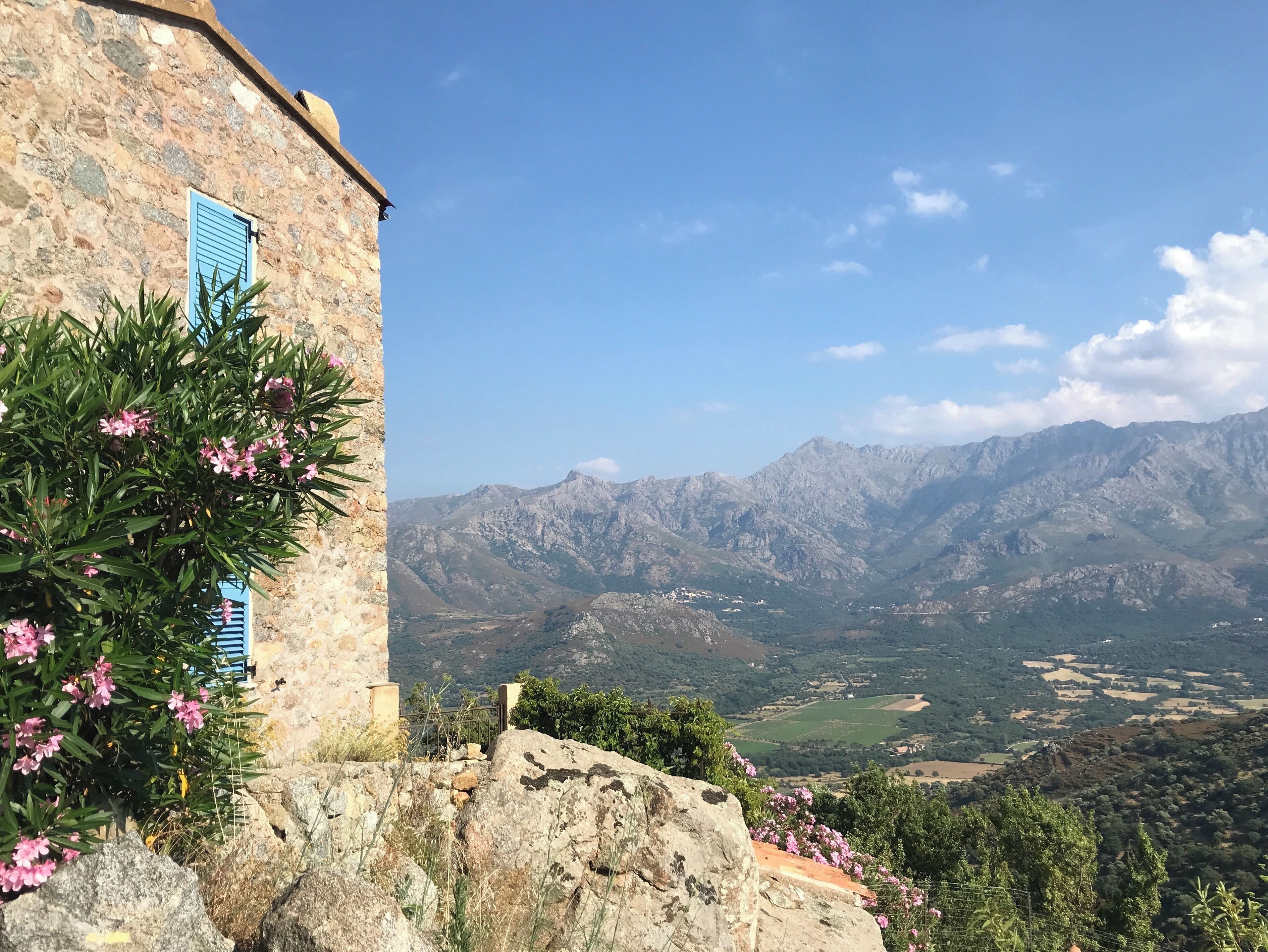 A stone building with blue shutters, pink flowers, and a mountainous landscape in the background.