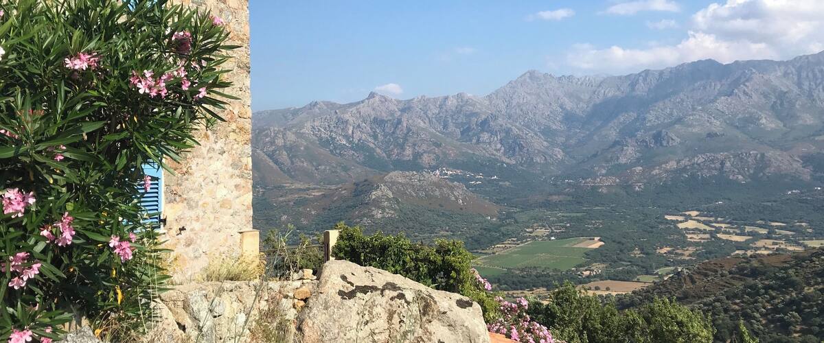A stone building with blue shutters, pink flowers, and a mountainous landscape in the background.