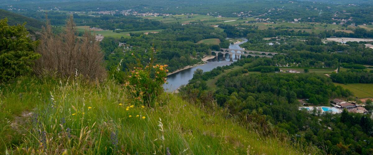 A river flowing through a valley with a bridge, surrounded by greenery and distant hills.