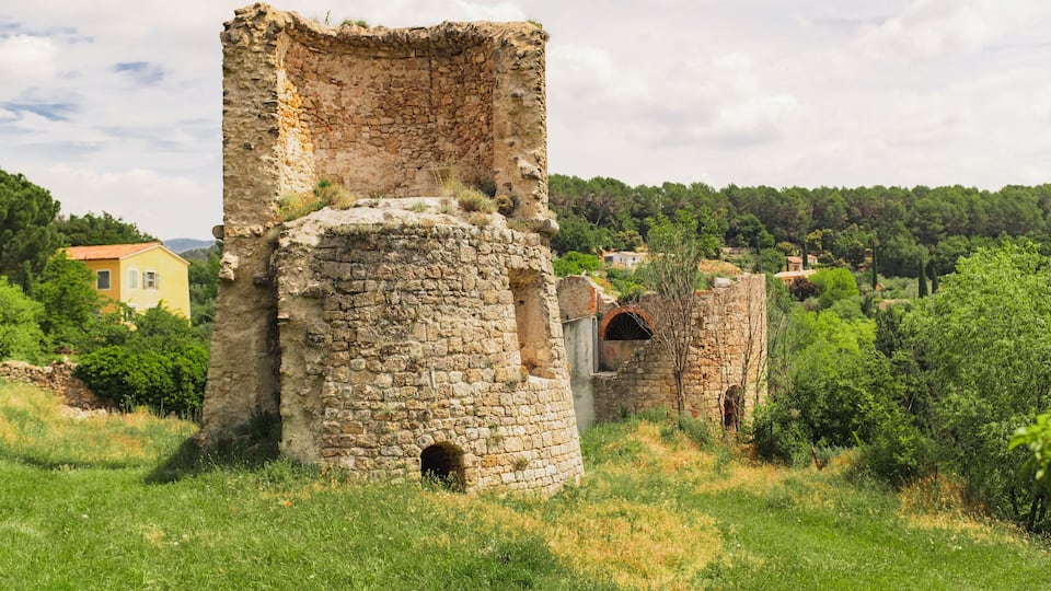 Ancient stone ruins with a circular tower and a rectangular building, surrounded by greenery and distant houses.
