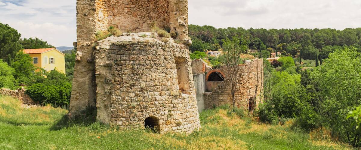 Ancient stone ruins with a circular tower and a rectangular building, surrounded by greenery and distant houses.