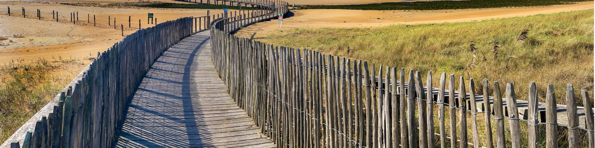 A wooden boardwalk curving through a coastal landscape with sandy dunes and grassy areas.
