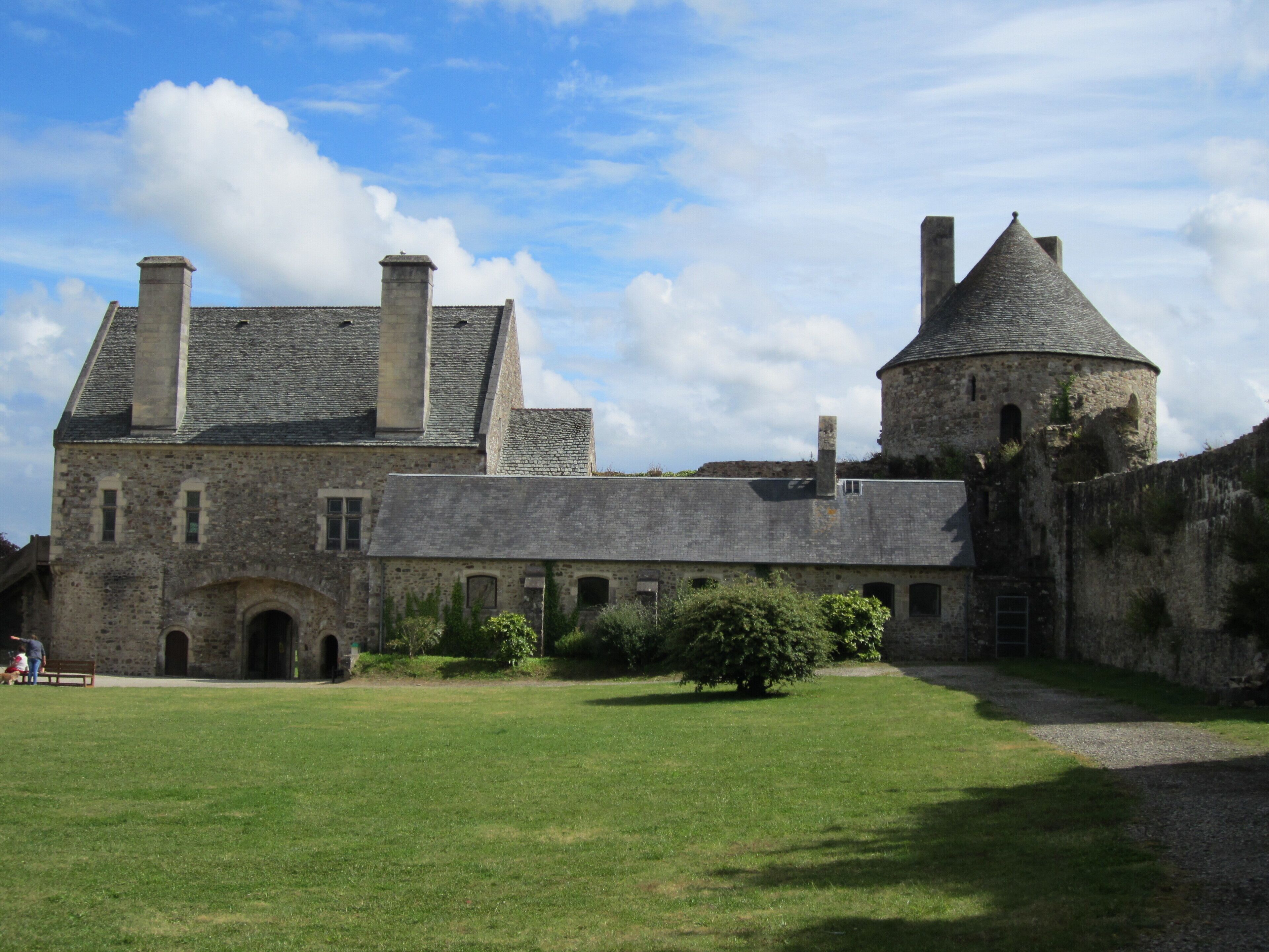 A historic stone building with a large courtyard, a round tower, and a grassy lawn.