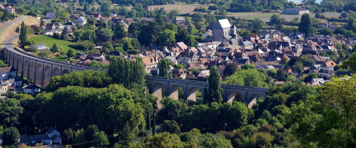 A town with a prominent stone bridge, lush greenery, and a river in the background.